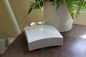 A compact Frontpoint security hub on a table near a plant, with indicator lights showing it is powered on.