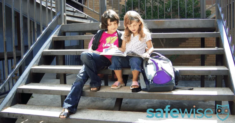 Two little girls with backpacks reading while waiting for the bus.